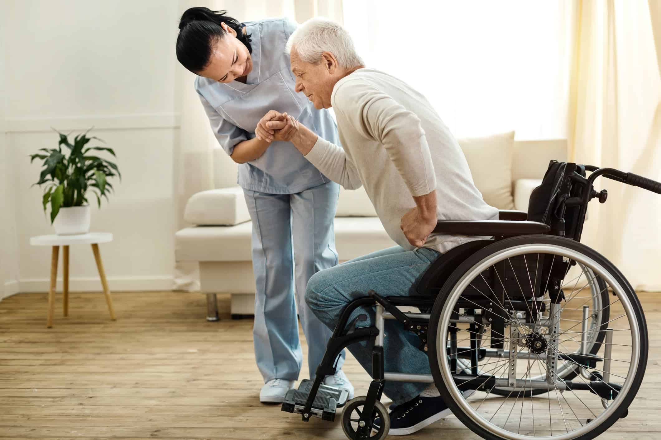 Caregiver helping an older adult stand beside a wheelchair at home